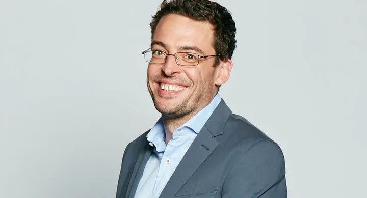 A man with glasses smiling, wearing a blue blazer and light blue shirt, stands against a plain gray background.