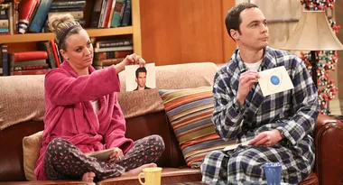 Two characters from "The Big Bang Theory" hold up different picture cards while sitting on a couch in pajamas.
