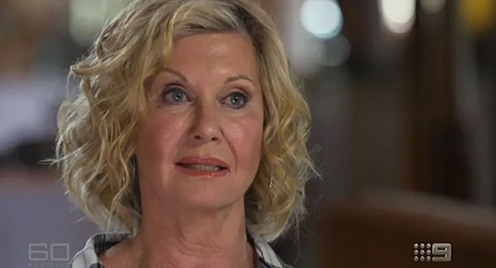 A woman with short, curly blonde hair speaks during a television interview, background slightly out of focus.