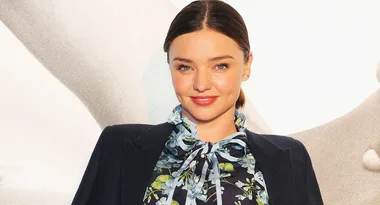 A woman with dark hair in a floral blouse and dark jacket smiles against a light background.
