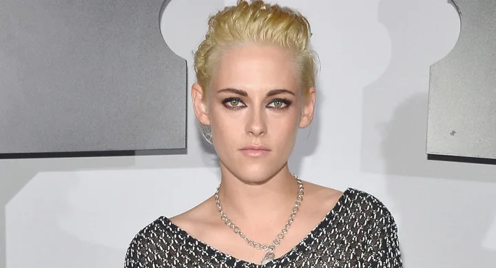 A person with short blonde hair wearing a patterned top and a chain necklace poses at an event.