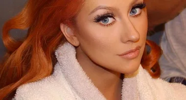 A person with red hair and blue eyes, wearing white, looks directly at the camera.