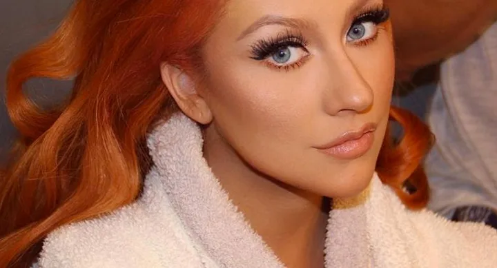 A person with red hair and blue eyes, wearing white, looks directly at the camera.