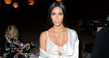 A woman in white lace and layered gold necklaces poses at an evening event with glowing lanterns in the background.