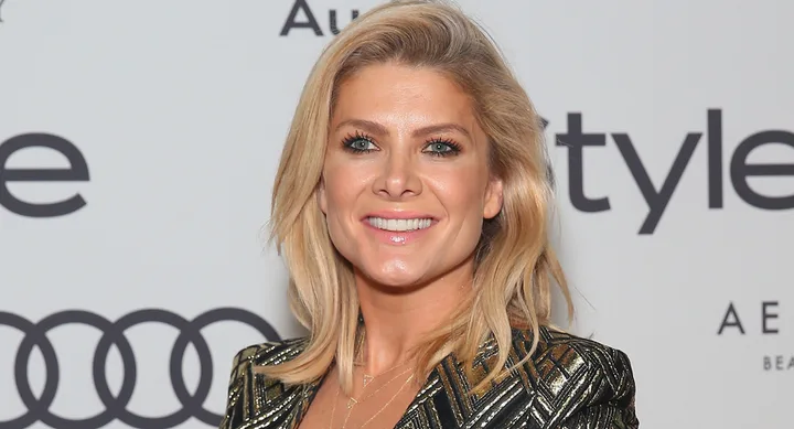 A person with blonde hair smiles at an event, wearing a patterned outfit with a backdrop featuring Audi and style logos.