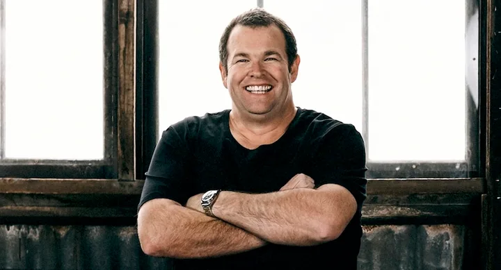 A smiling man in a black shirt with arms crossed, standing indoors in front of large windows.