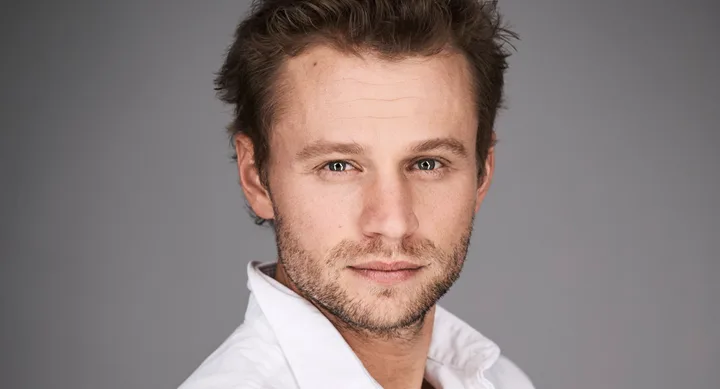 Man with short brown hair wearing a white shirt against a gray background.