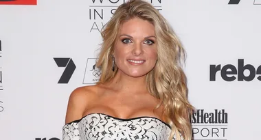 A person with long blonde hair smiles at an event, wearing an off-shoulder patterned dress against a backdrop with logos.