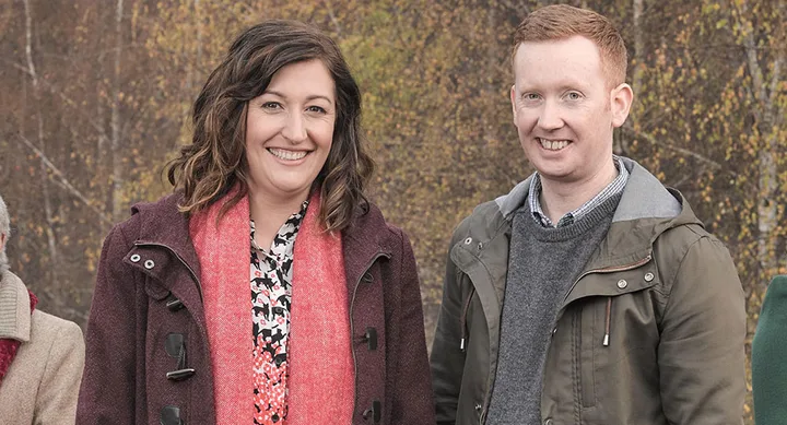 Two people smiling outdoors with autumn trees in the background, wearing coats and scarves.