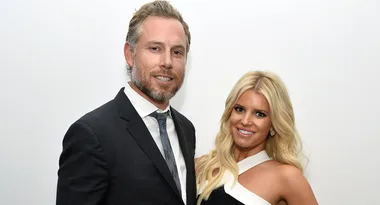 Man in a suit and woman in a white and black dress smiling together against a plain background.