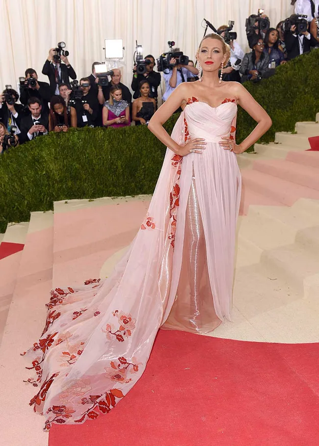 Woman in elegant pink gown with floral details posing on red carpet, surrounded by photographers.