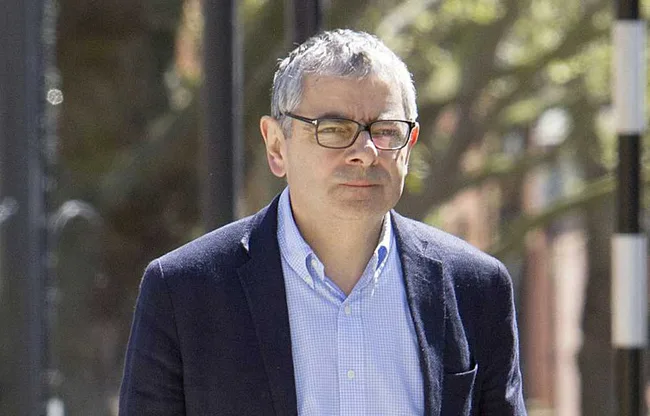 A man with glasses wearing a blue shirt and blazer walking outdoors on a sunny day.