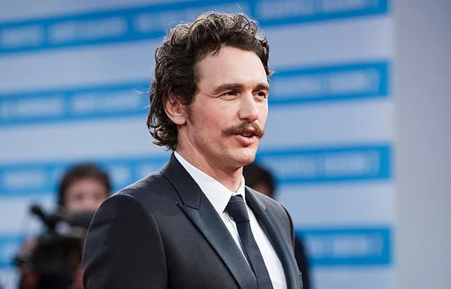 A man in a suit with a mustache stands on a red carpet, with a blue and white backdrop behind him.