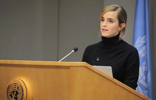 Woman speaking at a podium with a UN emblem, wearing a black top, and a blue flag is visible in the background.