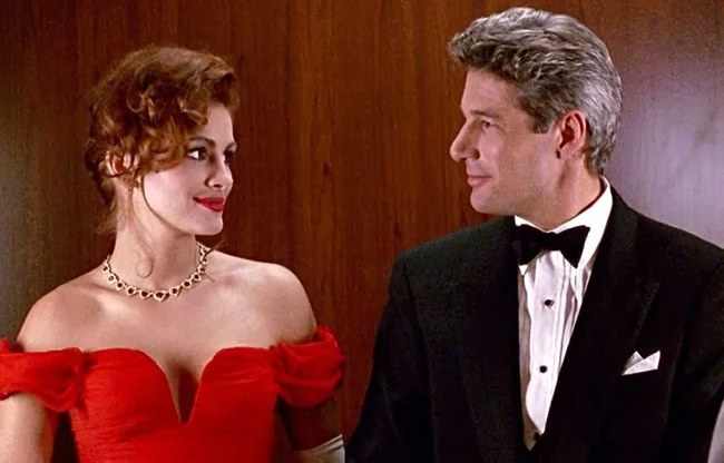 Pair in elegant attire, a woman in a red gown and a man in a tuxedo, stand smiling at each other against a wood-paneled background.