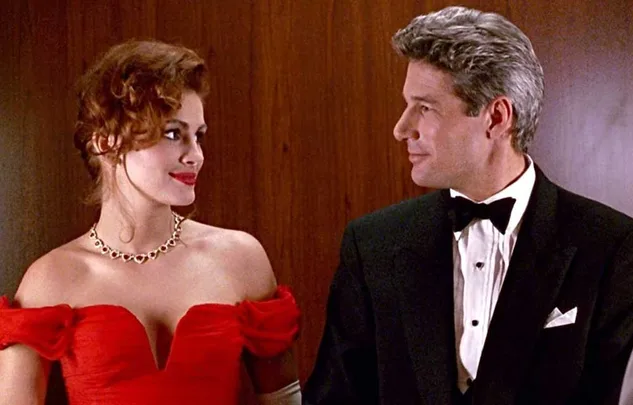 Pair in elegant attire, a woman in a red gown and a man in a tuxedo, stand smiling at each other against a wood-paneled background.