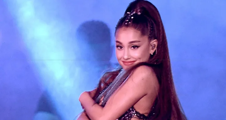 Singer on stage in a sparkly outfit, smiling with a blue and purple background.