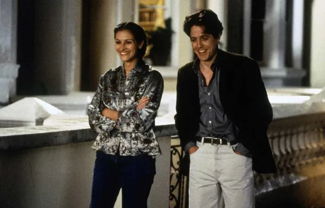 A woman and man casually smiling while walking on a terrace in a scene from a movie at night.
