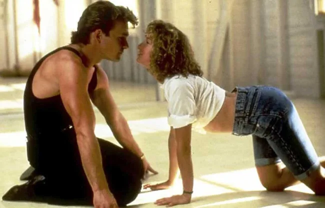 A man and woman kneel and face each other on a wooden dance floor, in a dramatic, intimate pose.