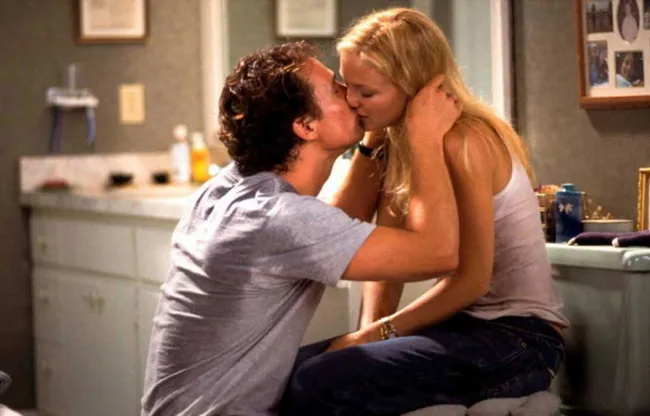 A couple kissing in a bathroom, with cabinets and a mirror in the background.