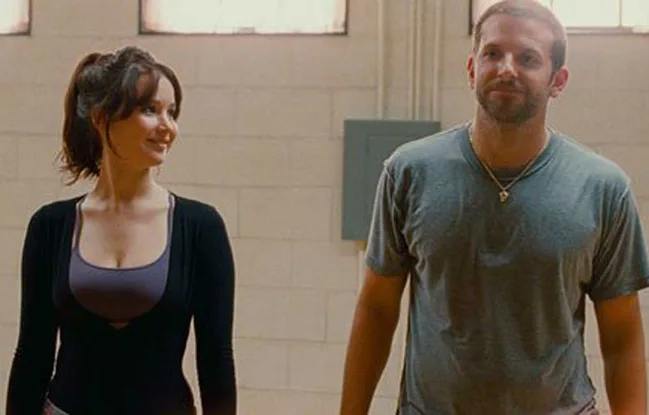 Two people standing side by side in a room, one smiling, both wearing casual clothing.