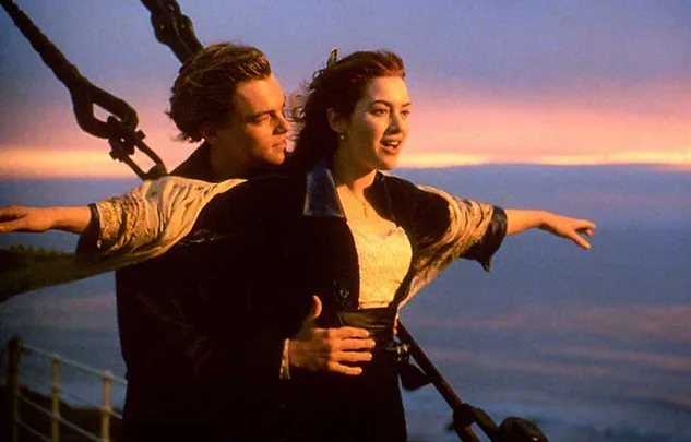 A couple embraces on a ship's deck at sunset, arms outstretched, with a view of the ocean.