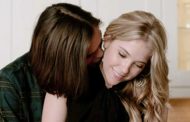 A person whispers to a woman who smiles with her eyes closed, showing affection and warmth in a light room.