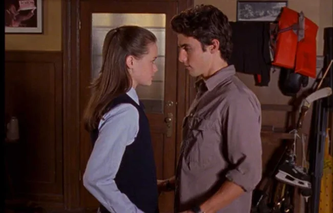 Two young people stand facing each other closely in a room with sports equipment and jackets on the wall.