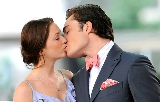 Two people kiss; a woman in a purple dress and a man in a suit with a pink bow tie in a blurred outdoor setting.