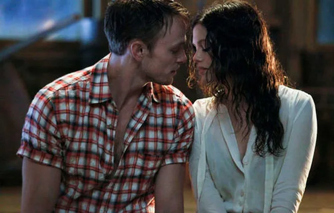 A man in a plaid shirt and a woman in a white blouse sit closely, appearing to share an intimate moment.