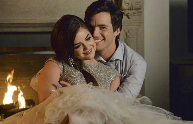 A couple sits smiling by a fireplace; the woman wears a sparkling dress and the man hugs her from behind.
