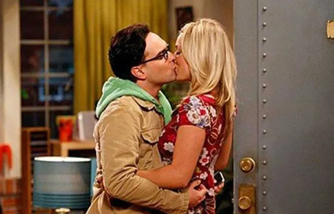 A man and woman share a kiss in a cozy room, standing near an open door with warm lighting in the background.