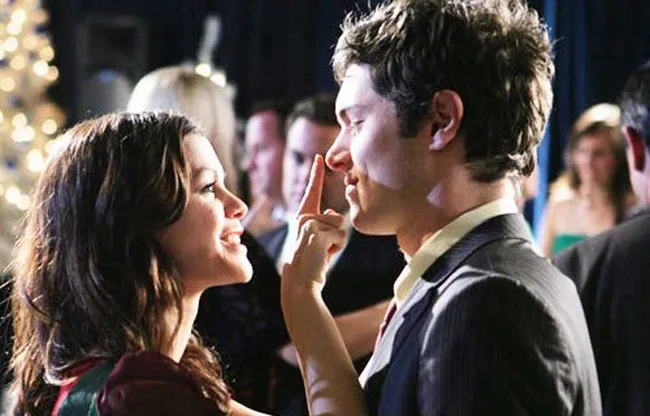 A woman playfully touches a man's nose at a festive party, with people blurred in the background.