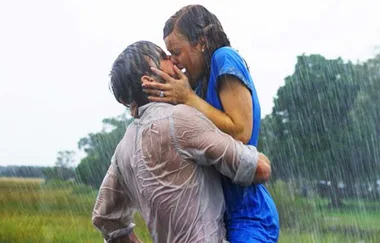 A couple kisses passionately in the rain, both wearing wet clothes in a lush outdoor setting.