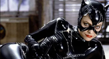 A person in a stitched, shiny black cat costume with red lipstick, looking at the camera in a dimly lit room.