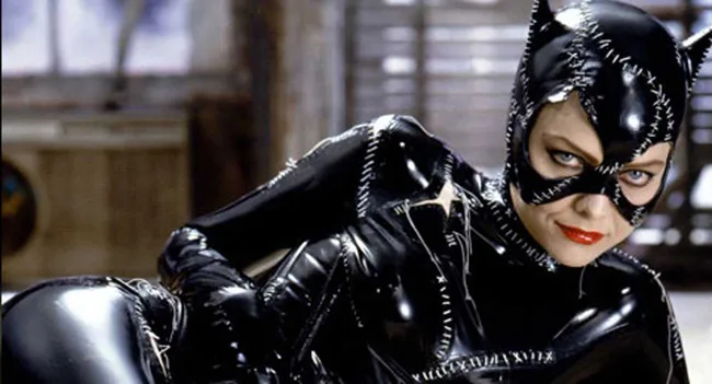 A person in a stitched, shiny black cat costume with red lipstick, looking at the camera in a dimly lit room.