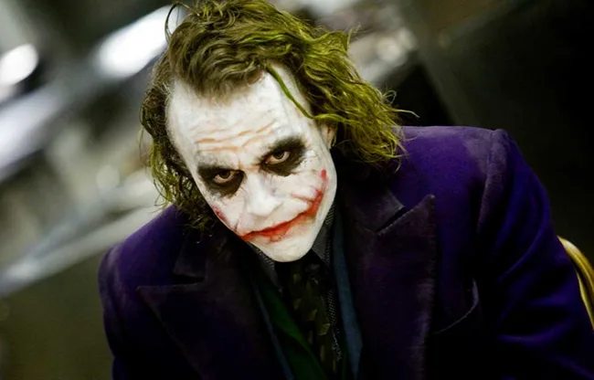 "The Joker character in makeup and purple suit, leaning forward with a menacing expression."
