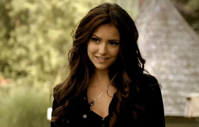 Young woman with long, wavy brown hair smiles while wearing a necklace and dark blouse, standing outdoors with blurred background.