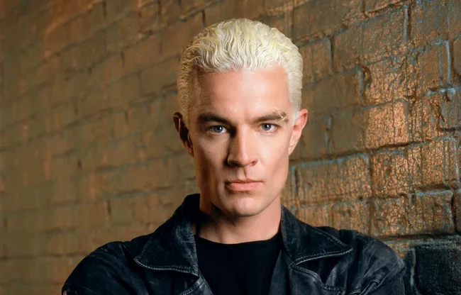 Young man with platinum blonde hair and black jacket poses against a brick wall, looking directly at the camera.