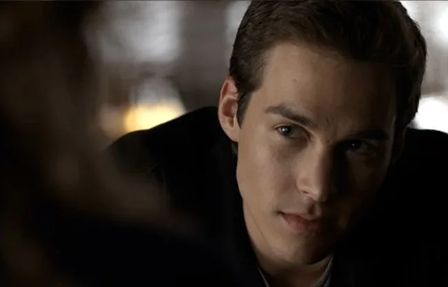 A man with short dark hair looks intently off-camera in a dimly lit setting.
