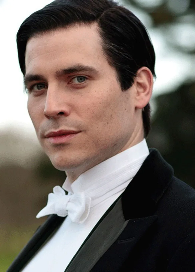 Man in formal attire with slicked back hair and a white bow tie, looking off-camera, set against a blurred outdoor background.