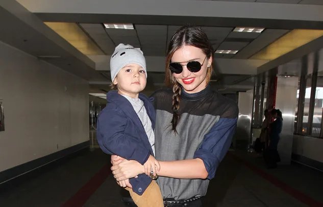 Woman in sunglasses holds a child wearing a grey beanie and blue jacket in an airport terminal.