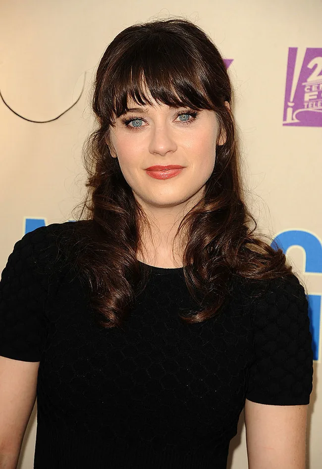 A woman with long dark hair and bangs, wearing a black top, stands against a neutral backdrop with a partial logo visible.