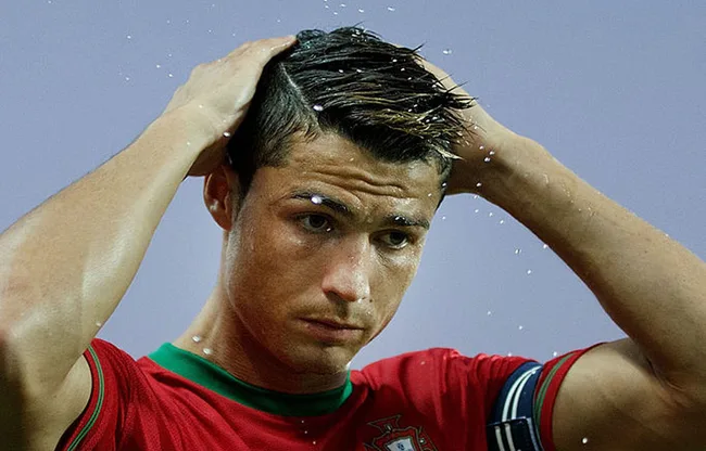 A soccer player in a red jersey adjusting his hair, with water droplets visible.