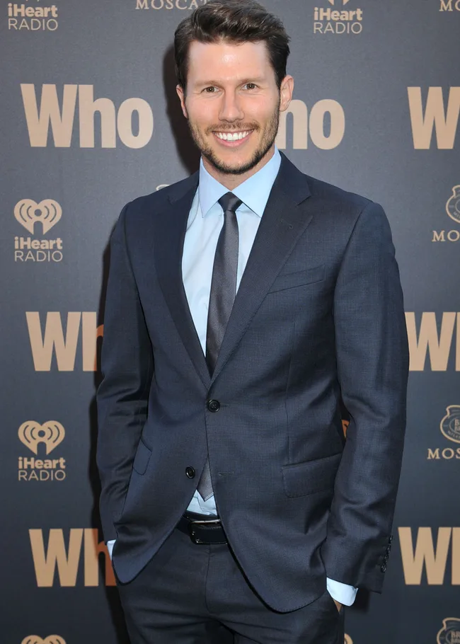 A man in a blue suit and tie smiles at a red carpet event with iHeart Radio and "Who" logos in the background.