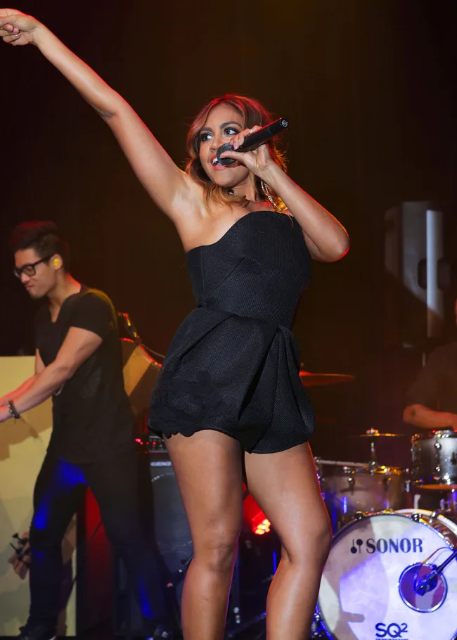 Singer performs on stage, wearing a black outfit, holding a microphone, with a band playing in the background.