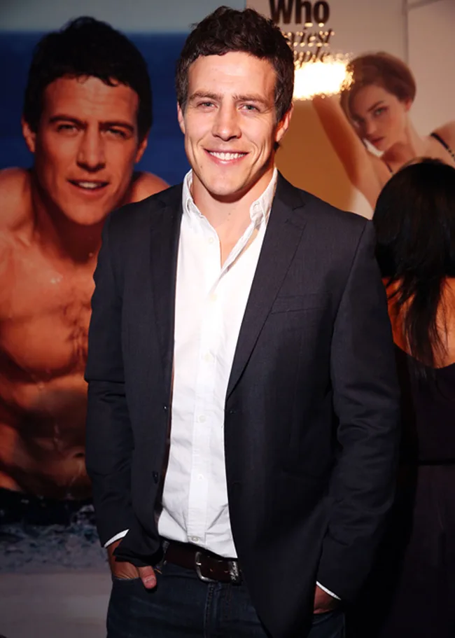 A person in a dark blazer and white shirt smiles at an event, with a beach-themed poster in the background.