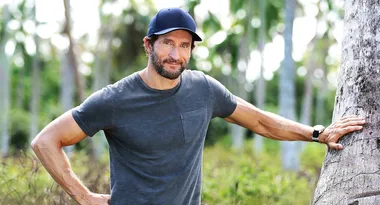 Man in casual shirt and cap leaning against a palm tree in a tropical setting.