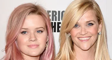 Two women smiling at a public event; one has pinkish hair, the other has blonde hair, both wearing earrings.