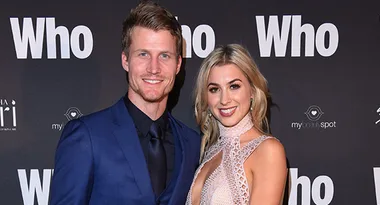 Man in a blue suit and woman in a lace dress posing at an event with "Who" logo backdrop.